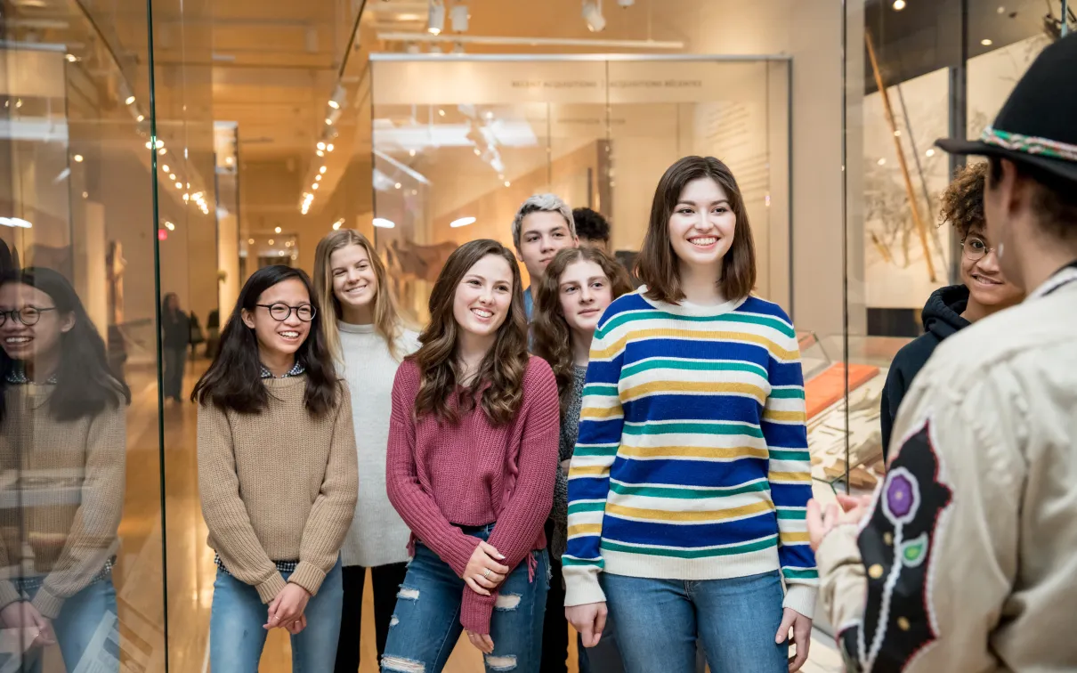 A diverse group of teenage students stands between cases in the First Peoples gallery and smile as they listen to an Indigenous Educator