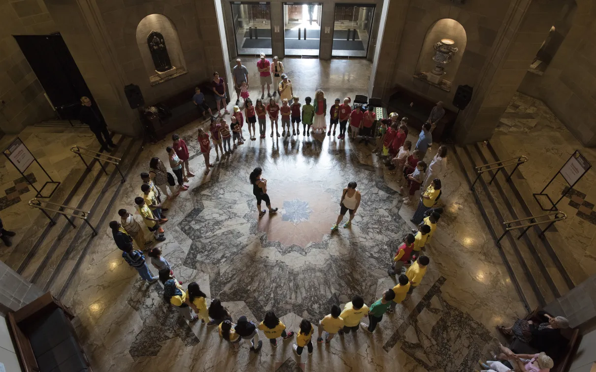 The camera looks down from above at group of students sitting in a circle at the bottom of a round ornate room