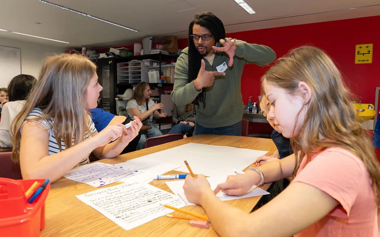Two students sitting at a table work on a plan for a porcupine quill stitching game. Another student looks up at an adult making a square shape with his fingers.