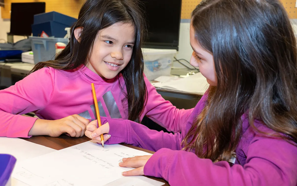 Two smiling young students look at each other. One is holding a pencil and writing.