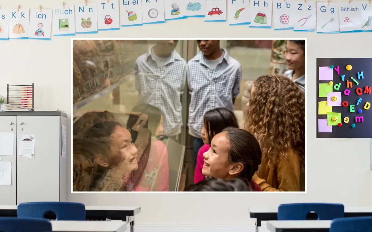 A screen in a classroom shows a scene of smiling students gathered around a museum case holding a mummified Ancient Egyptian