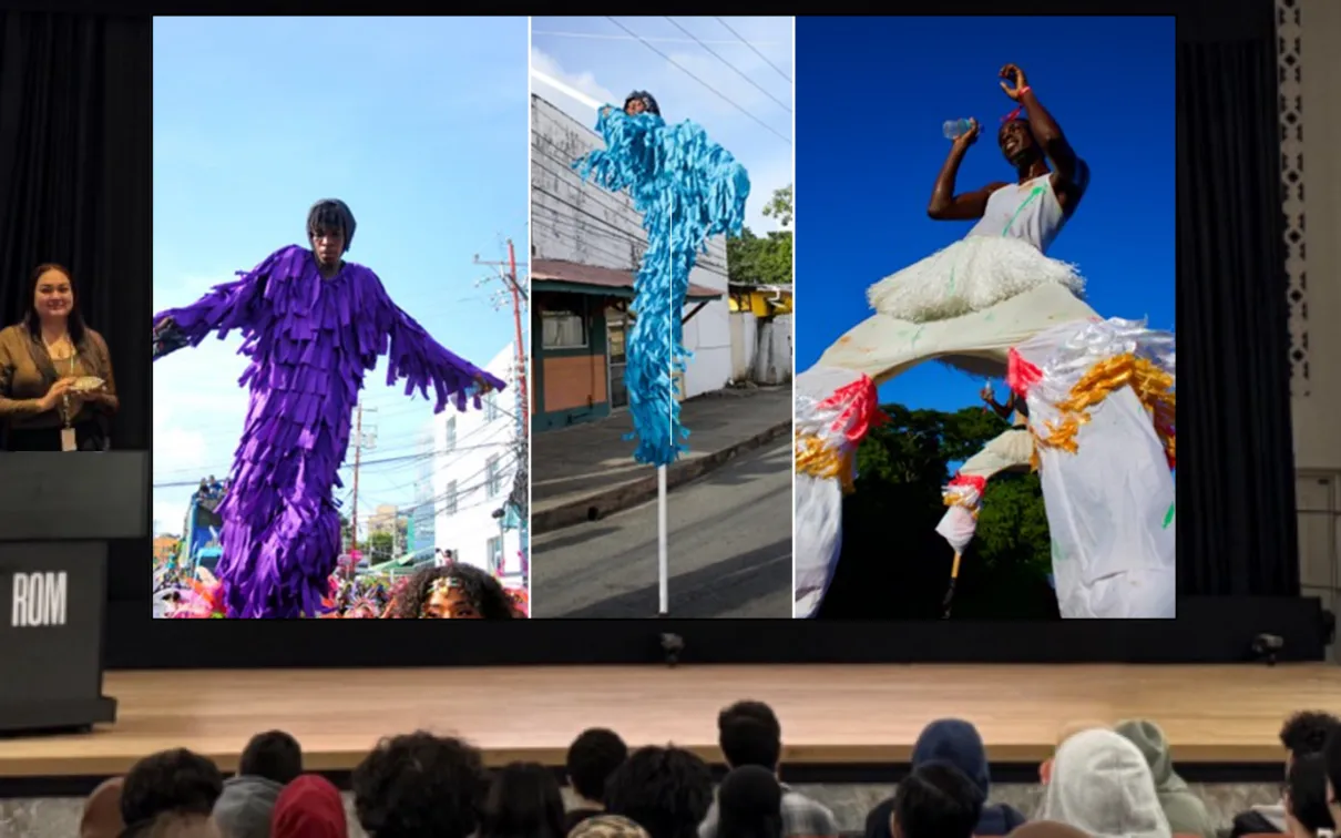 A presenter on a stage in front of an audience stands behind a podium labelled ROM. The screen next to them displays a composite image of three dark-skinned people wearing colourful fringed outfits and walking on stilts.