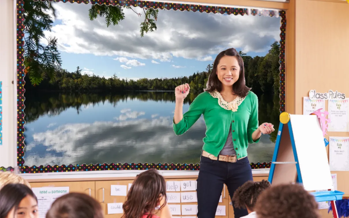 A teacher in a green cardigan stands in front of a smartboard at the front of a class. The board displays an image of a glassy lake.