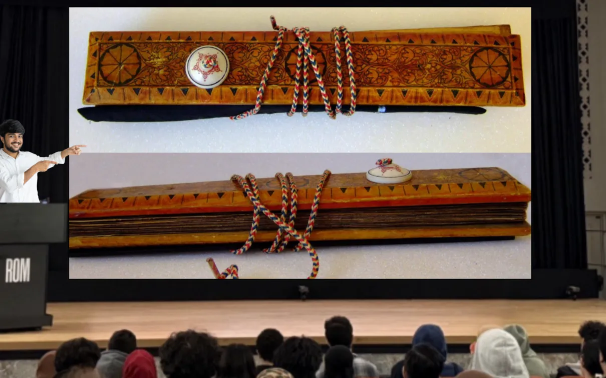 A smiling man stands behind a podium labelled ROM on a stage and gestures at a screen as an audience watches. The screen displays two images of a manuscript held between two ornately decorated wooden covers.