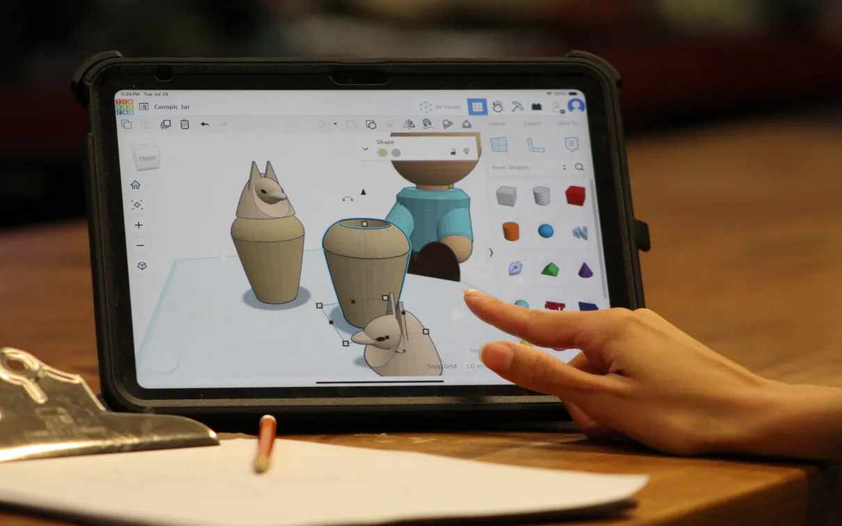 A hand touches an ipad displaying 3D models of Egyptian objects