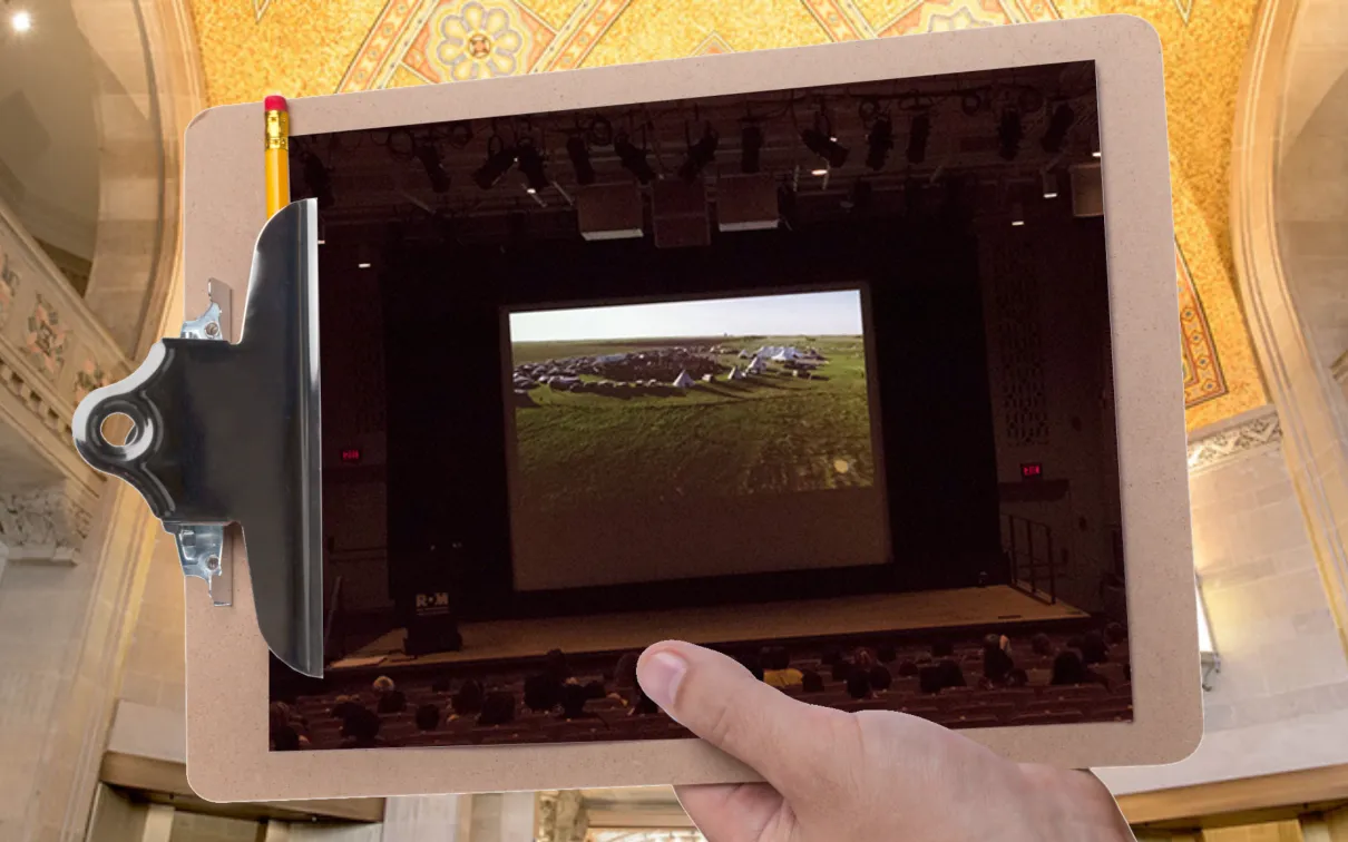 A hand holds a clipboard in front of a large gallery with a gold mosaic ceiling. The clipboard displays a picture of students in a darkened theatre watching a presentation.