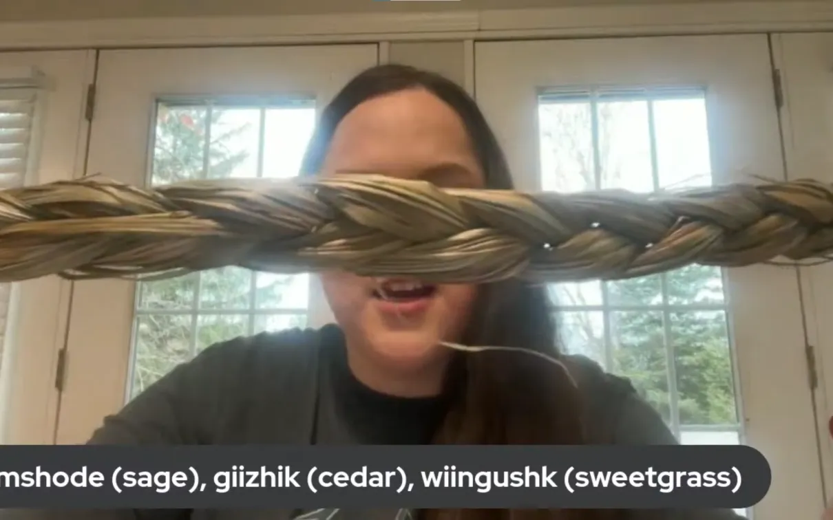 An Indigenous woman holds up braided sweetgrass. A caption on the bottom of the screen reads mshode (sage{, giishik (cedar), wiingushk (sweetgrass)