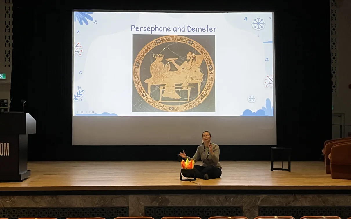 A woman sits on a theatre stage next to an artificial campfire. A screen behind her shows a slide titled Persephone and Demeter, with an image of two goddesses depicted in red-figure Greek pottery.