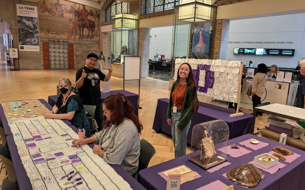 A station in a large gallery covered in activities, objects, and a white wampum belt with a purple circle at the back. Staff man the activities.