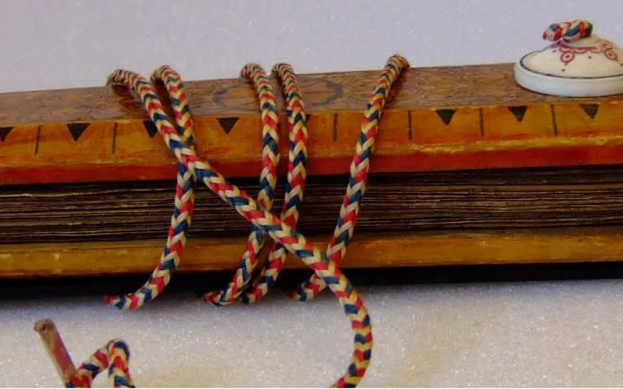 A side view of a manuscript sandwiched between two wooden cases. Decorations visible on the top case include black circles, and alternating lines and triangles. A white button on the upper case secures a red, white, and blue cord that wraps around both cases and holds the manuscript together.