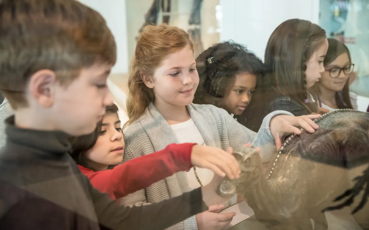 Several young students in a diverse class touching a bronze statue of a maiasaur.
