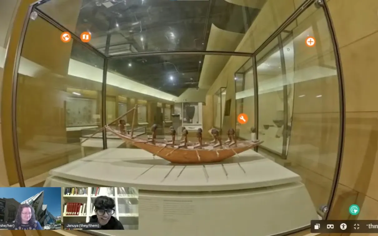 A display showing a wooden model of a boat filled with small humanoid figures with black skin and dark hair. Facecam inserts of two smiling people are at the bottom of the screen.