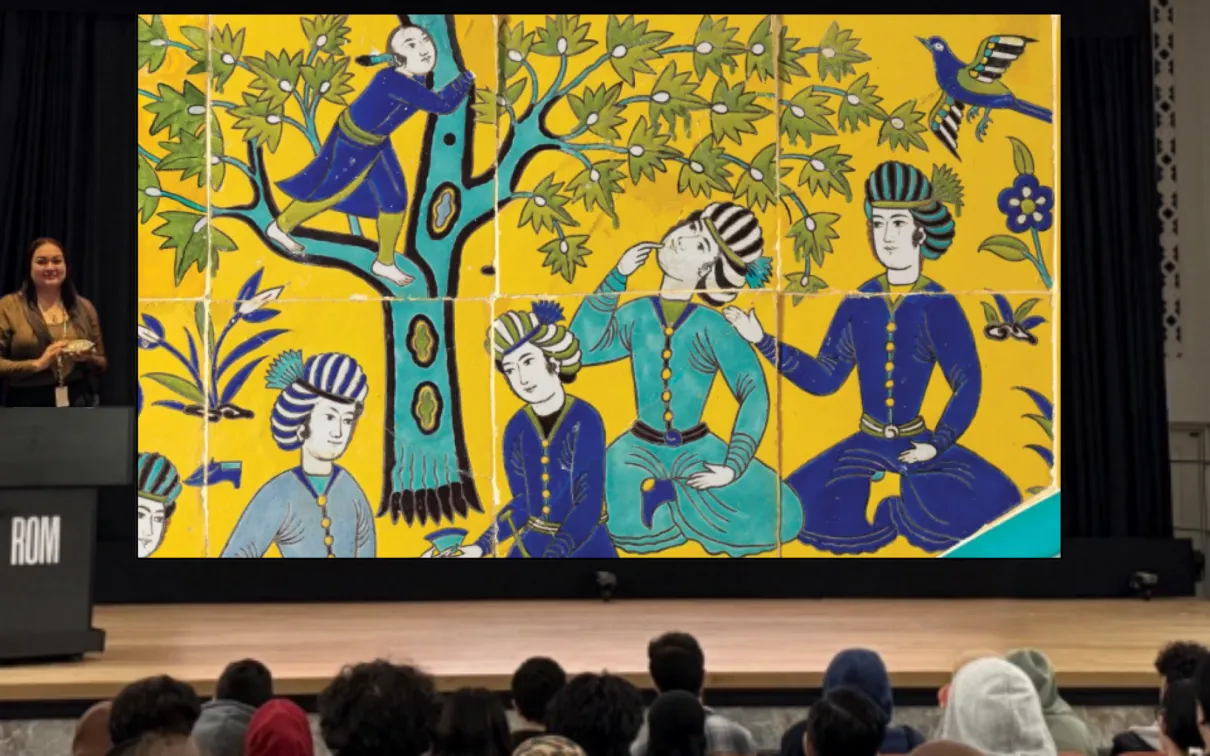 A woman stands behind a podium on a stage. The screen behind her displays an image of a colourful Islamic tile depicting several people at a picnic.