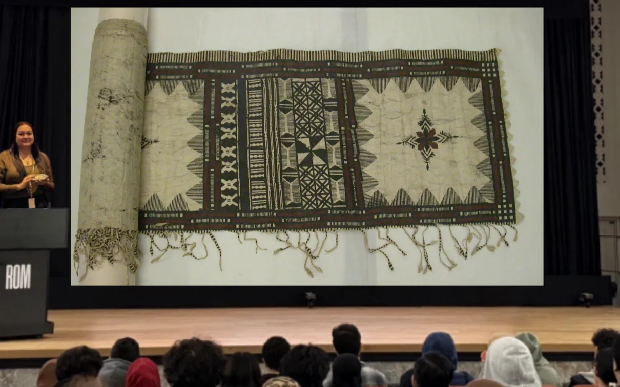 A smiling Indigenous woman stands behind a podium on a stage. On a screen next to her is an image of a length of cloth with intricate patterns in dark black and brown ink.