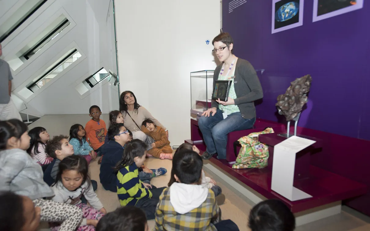 Several young students in a diverse class seated in front of an Educator who is showing them images on a tablet.