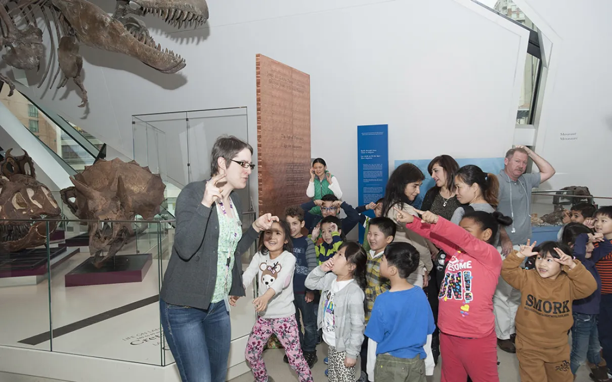 Several young students in a diverse class using fingers to make horns on their foreheads and pointing at teacher who is making claws with her fingers.