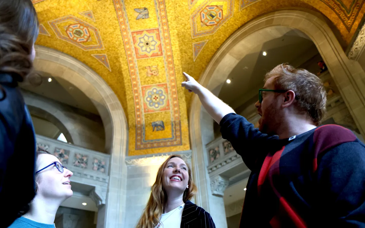 Three happy people look up at an ornate golden ceiling as one person points out a detail.