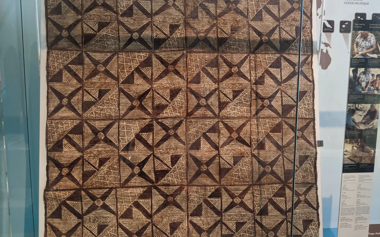 A large square of brown and beige cloth hangs in a museum display case. The cloth has intricate geometric patterns made up of lines, squares, and triangles.