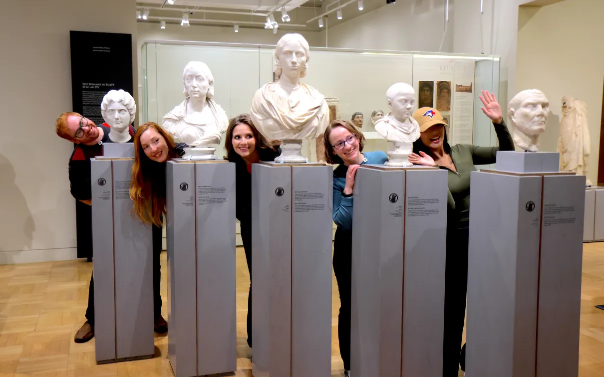 “A group of people playfully pose by leaning out between classical marble busts in a museum gallery.