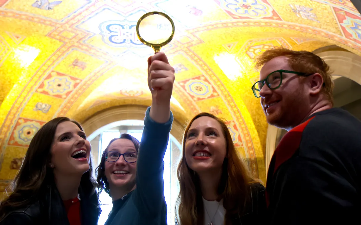 A group of people smile and hold up a magnifying glass while exploring the ornate golden ceiling of the museum rotunda.