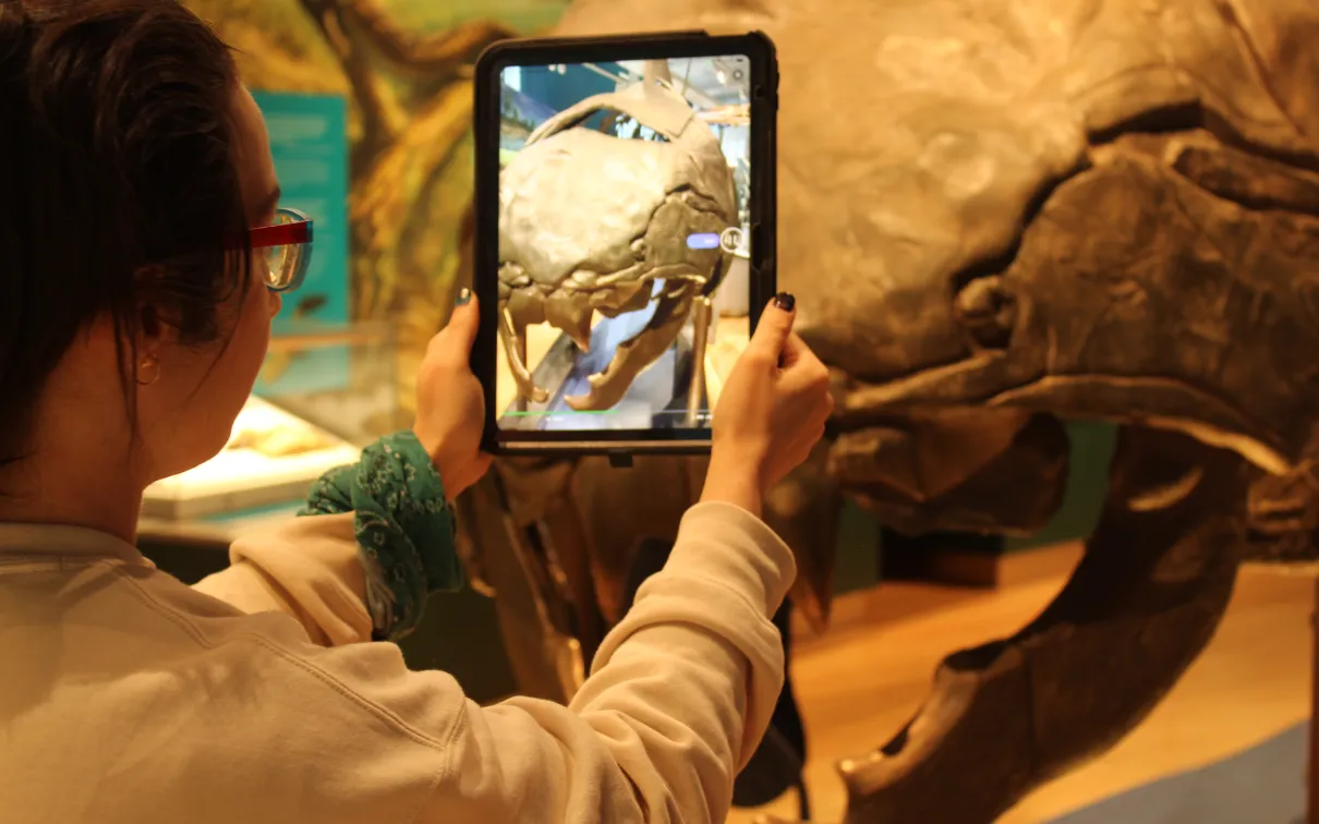 A person stands in front of a large fossil fish head holding an ipad that displays the head from the front.