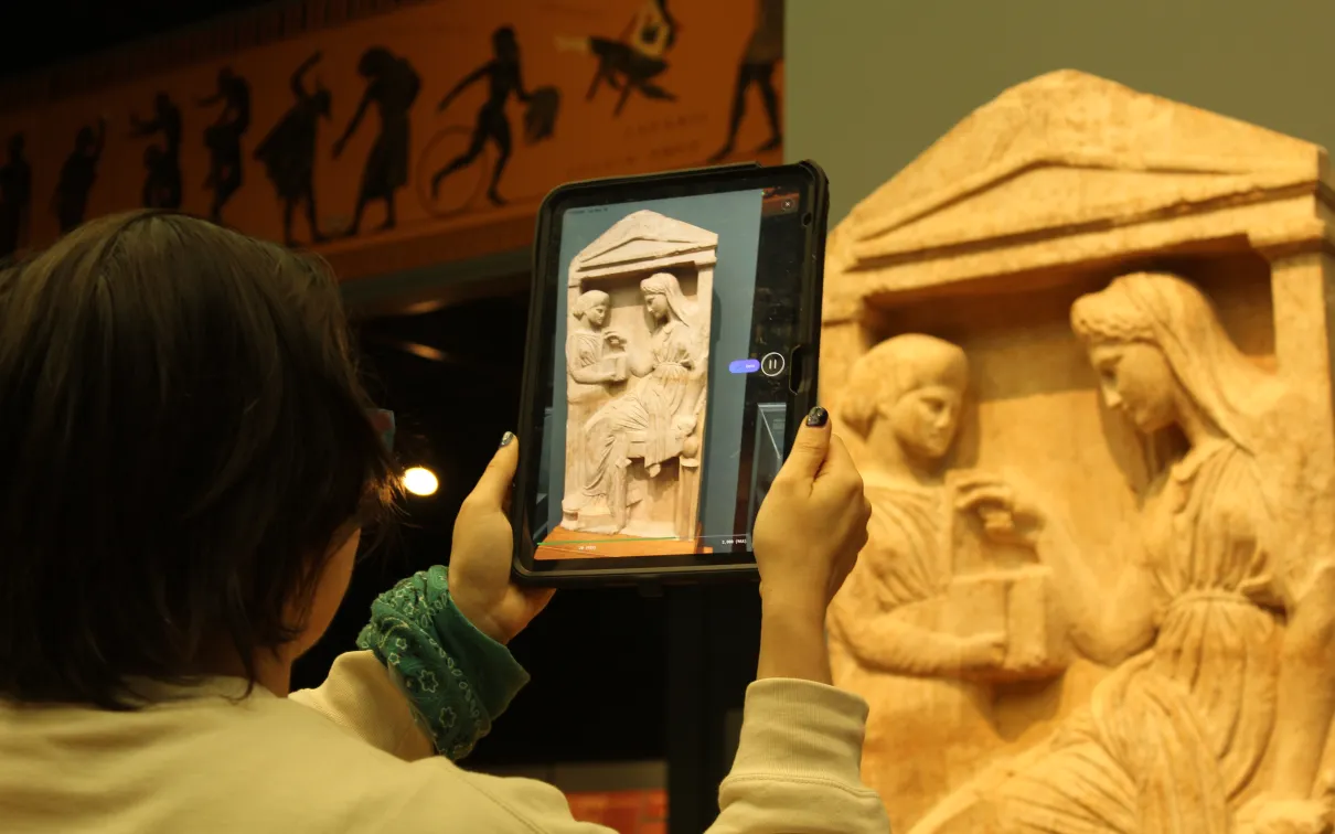 A person holds a tablet up to scan a Greek carving of a woman choosing jewellery from a box.