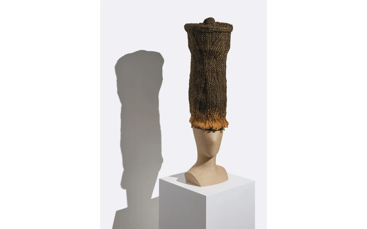 Tall cylindrical woven basket with a flared rim displayed on a white pedestal casting a shadow on the wall behind.