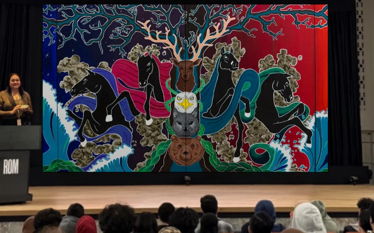 An Indigenous woman stands on a stage behind a podium. A screen behind her displays a painted mural.