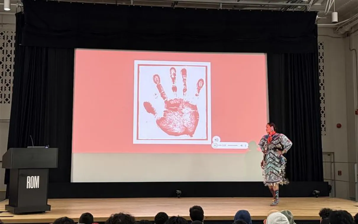 An Indigenous woman wearing a jingle dress stands on a stage in a theatre. The screen behind her shows a white handprint on a red screen.