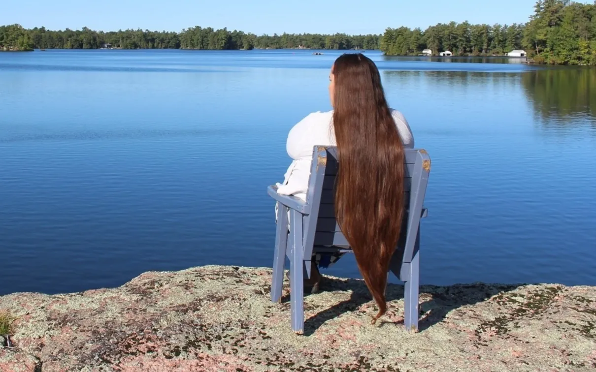 A woman with long hair sits on a chair overlooking a glassy blue lake.