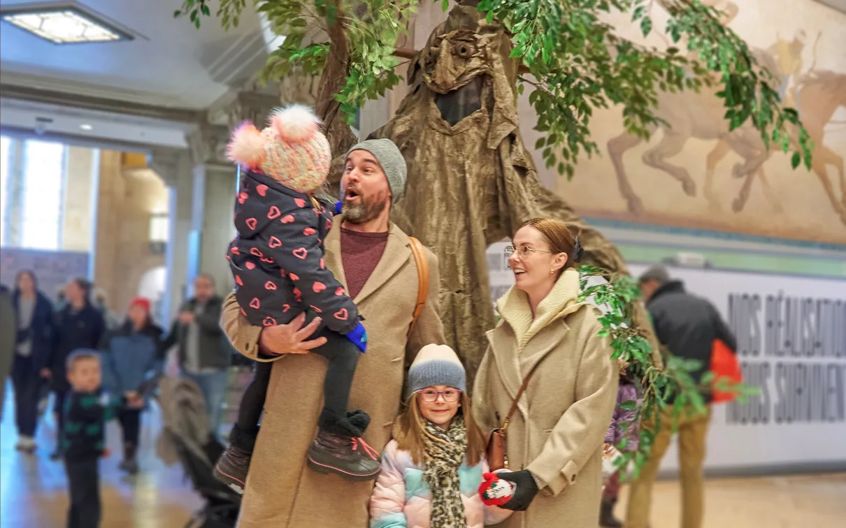 Two adults and two children stand indoors near a large tree-like display; one adult holds a bundled-up toddler while the others smile, with people walking in the background.