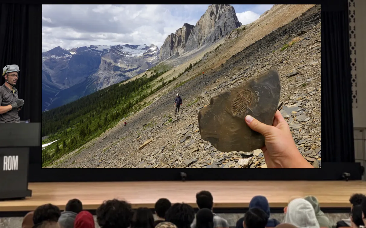 A man wearing a hard hat with goggles stands behind a podium on a stage. A screen next to him depicts a shale-covered hillside with mountains in the background. A lone figure walks across the hillside. A hand holds up a fossil in the foreground.