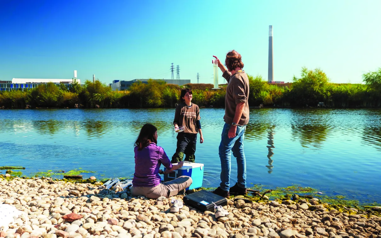 Three people conduct water sampling along a rocky shoreline, with one person holding up a clear measuring tube while others handle containers and equipment beside a river; trees, industrial buildings, and tall smokestacks are visible across the water under a clear blue sky.