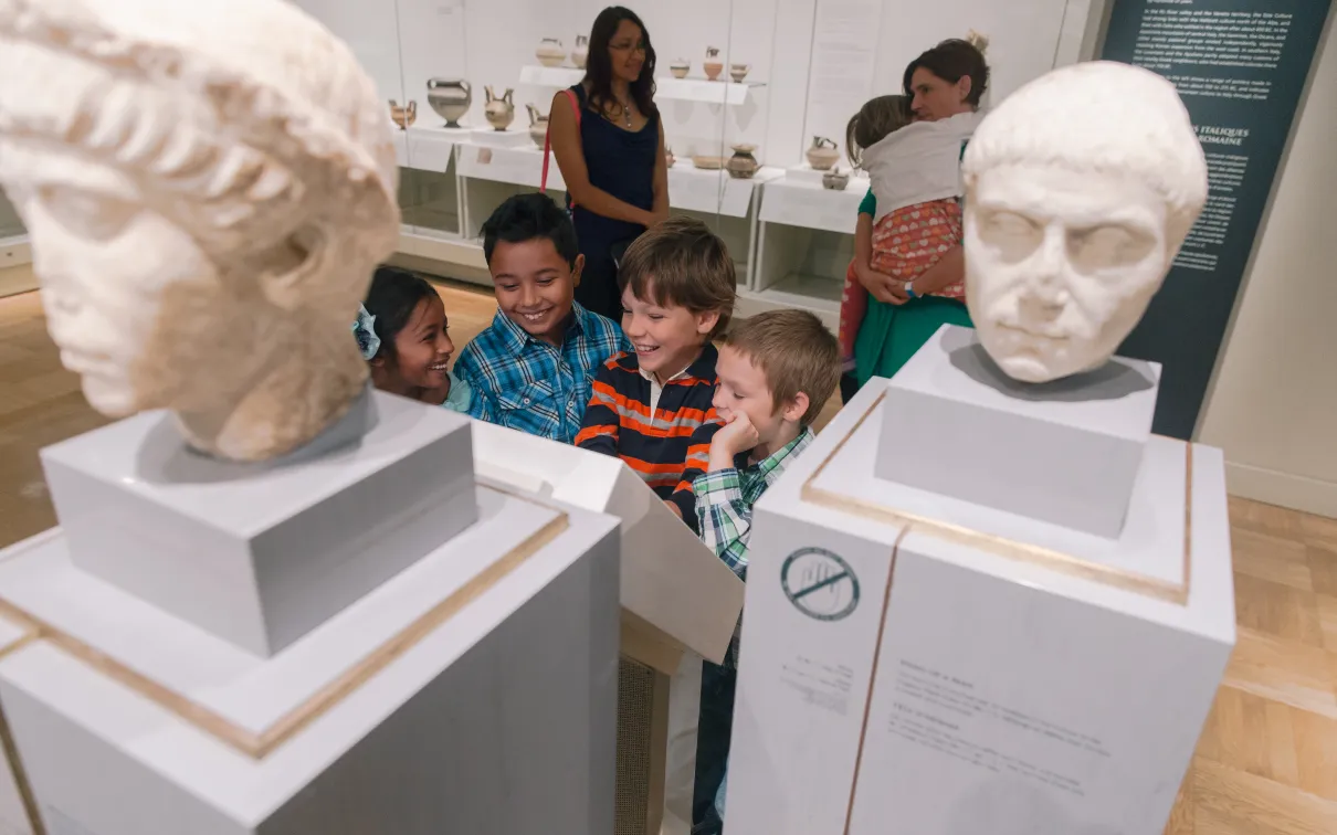 Two adults watch a smiling group of children in front of a group of marble busts