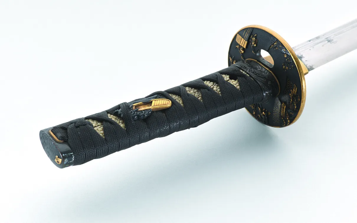 Close-up view of a Japanese sword hilt with black cord-wrapped handle, textured white ray-skin beneath, and ornate black-and-gold guard, photographed diagonally on a white background with soft lighting.