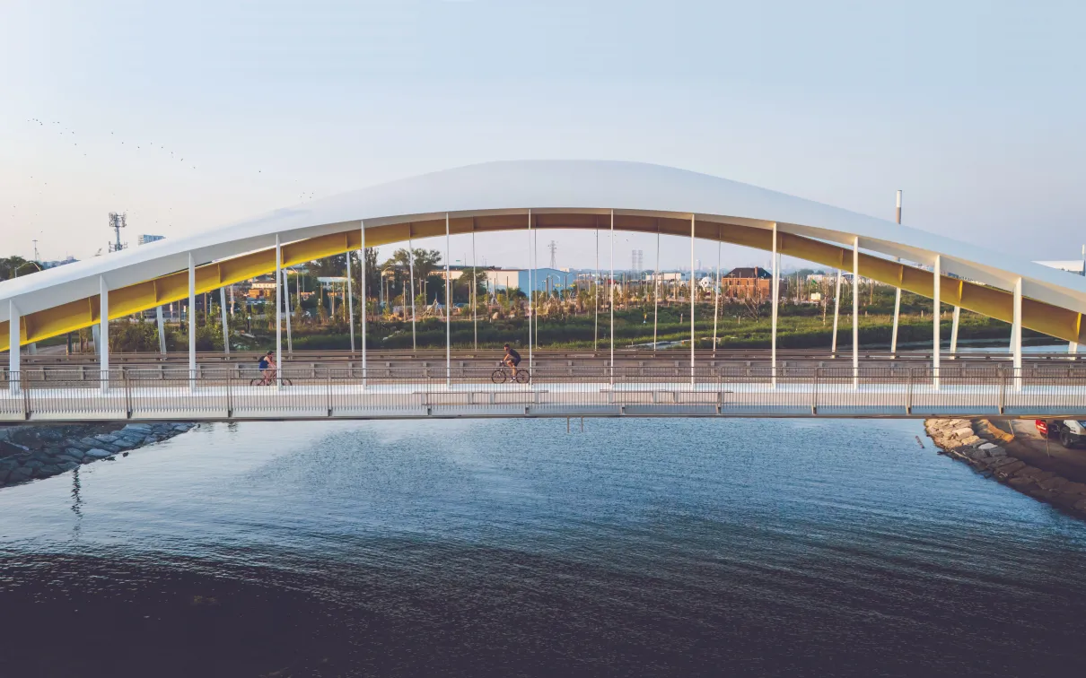 Wide view of a modern arched pedestrian bridge spanning a body of water, with cyclists crossing beneath a curved white canopy supported by vertical cables, set against a city landscape and pale blue sky in warm evening light.