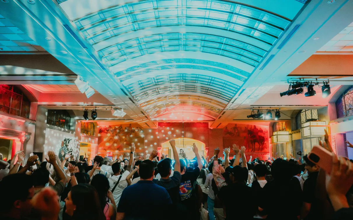Crowd of people in a large indoor venue with a high curved glass ceiling, raising their arms toward a brightly lit stage. Colorful red and blue lighting and spotlights illuminate the space, with artwork and event lighting visible along the walls