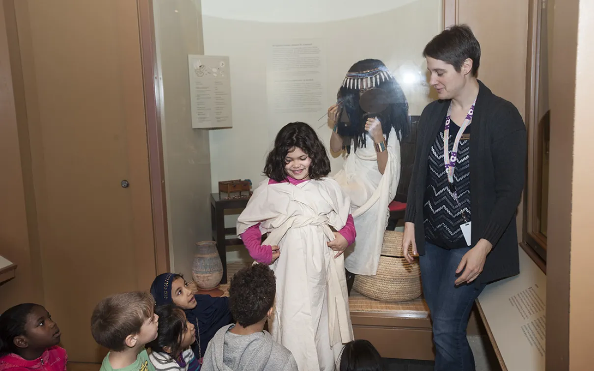 A museum teacher dresses a smiling child in an Egyptian dress as classmates watch.