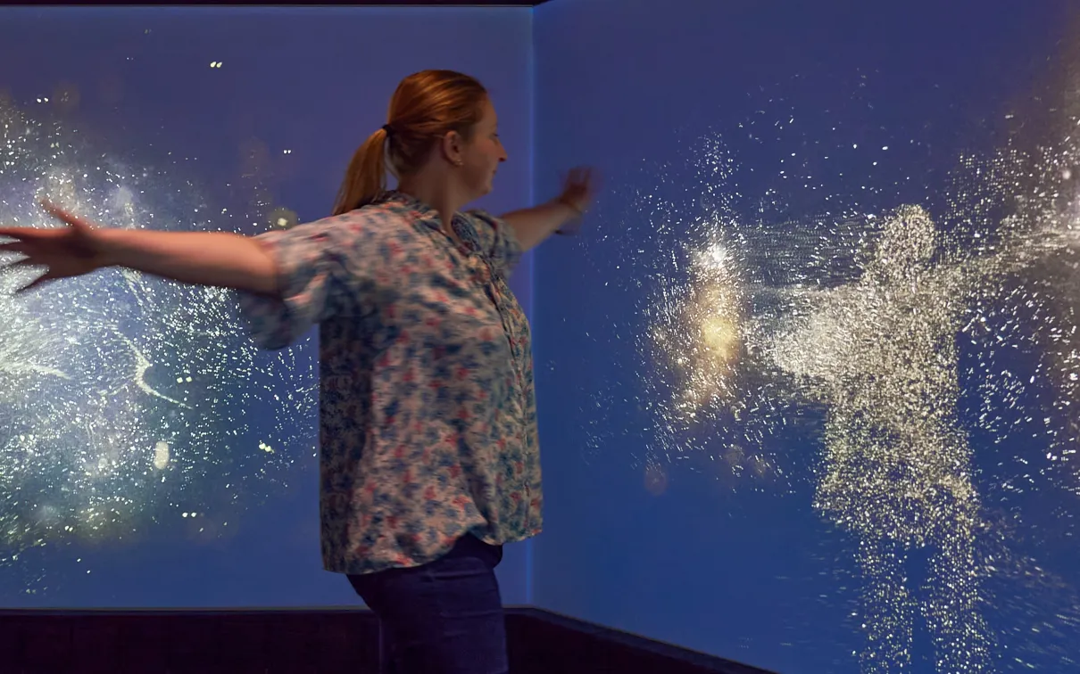 A woman moves in front of an interactive screen where clusters of pollen-like light follow her movements.