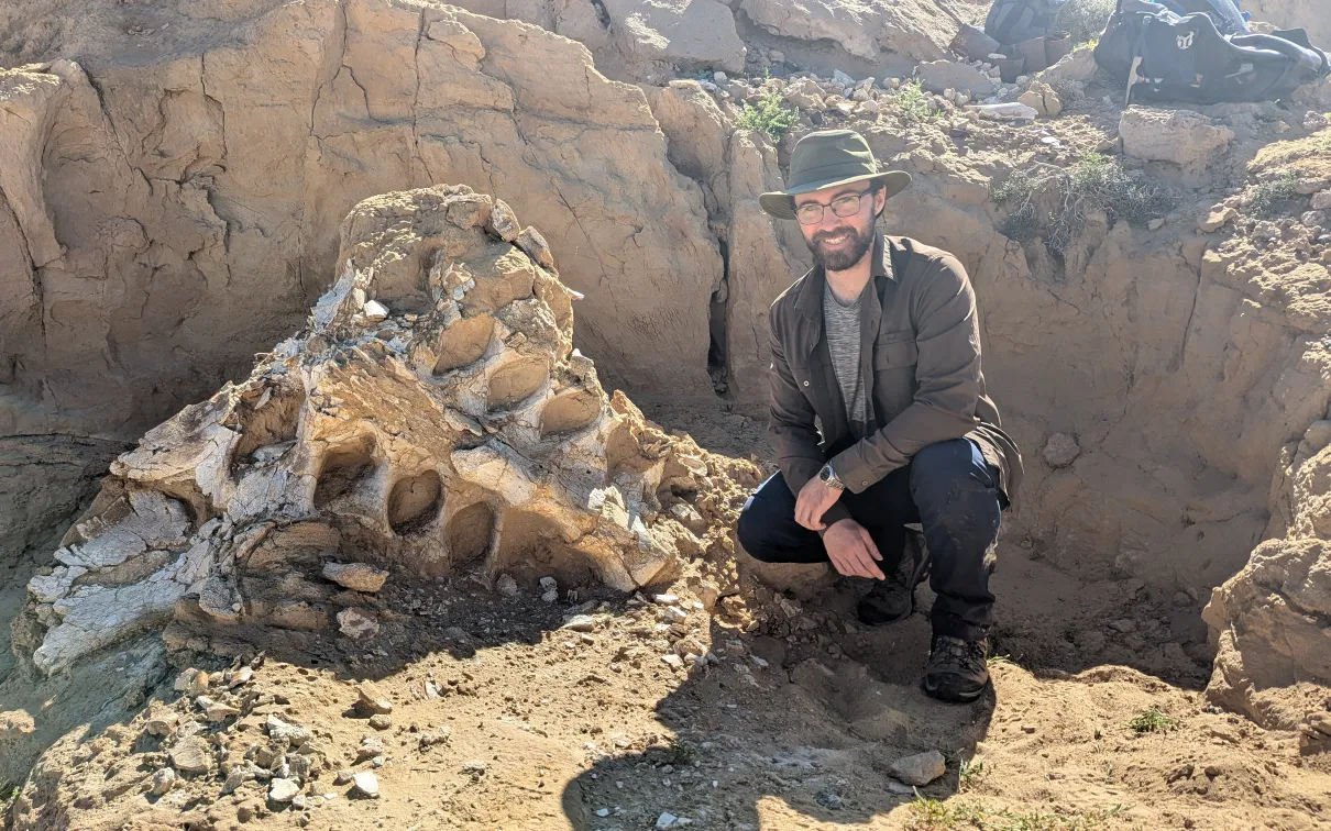 Tom Dudgeson wearing a baseball cap, sunglasses, and dark outdoor clothing crouches beside a large dinosaur skull partially embedded in rocky terrain.