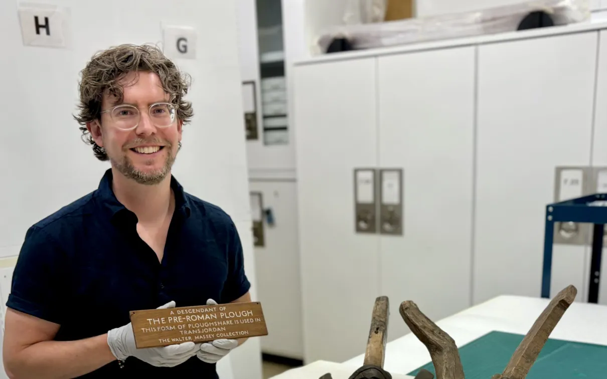 Greg Fester holding a wooden label that reads "MANITOBA - Plains Indian hide scraper," with three large hide scrapers on a table.