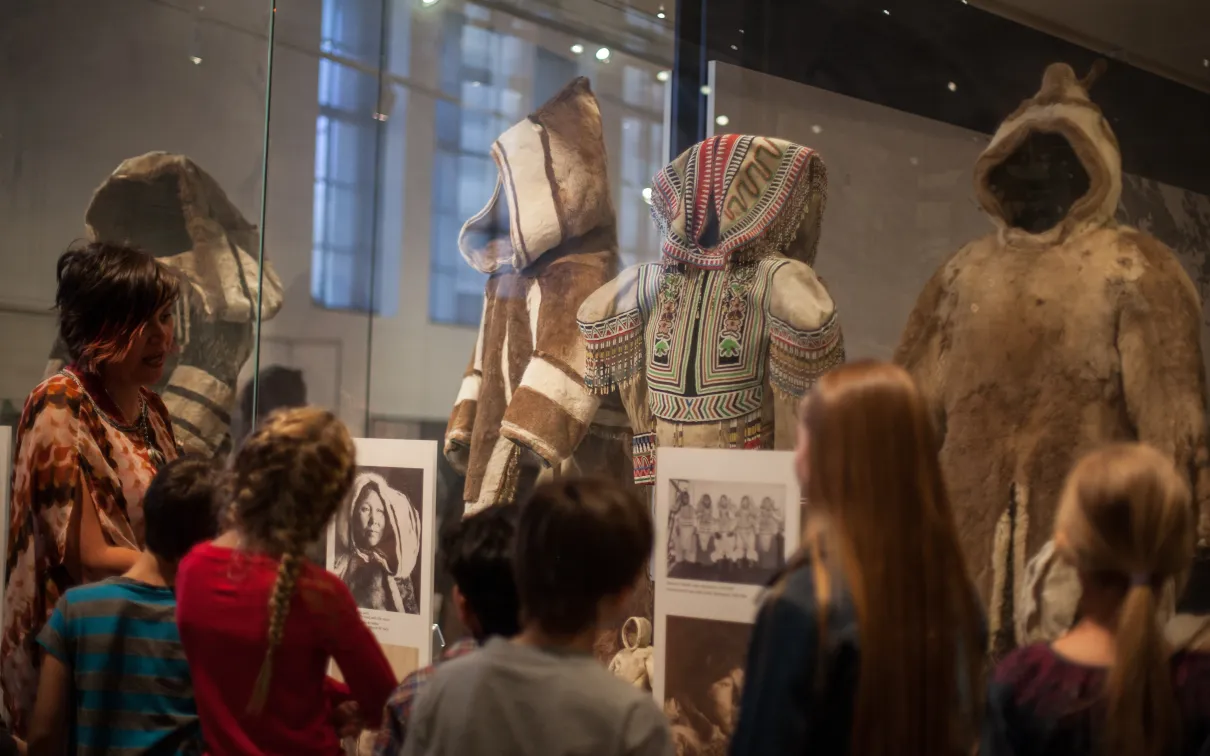 A teacher speaks to a group of students in front of a display case of sealskin parkas.