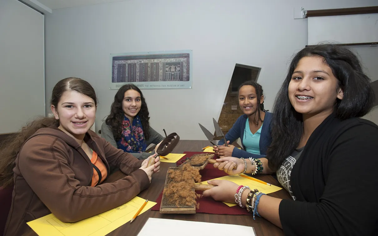 Four smiling students are looking at the camera sitting around a table holding a different tool.  One is holding a curry comb, one holds carding combs, and one is holding sheep shears.  