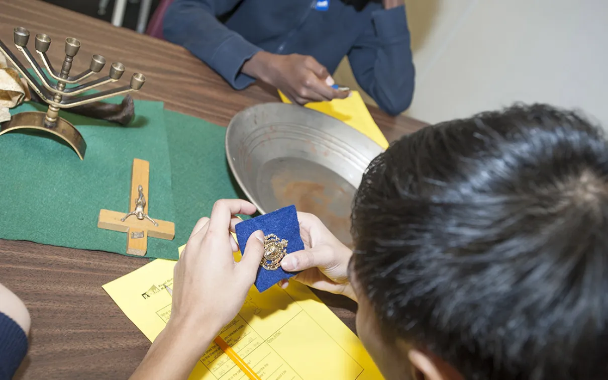 A student is holding a pin, while on the table in front of them is a menorah, a crucifix, and a tray for panning for gold. 