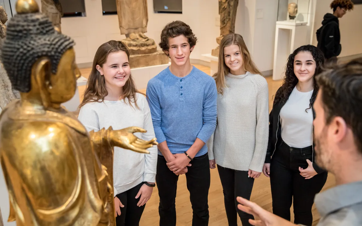 Four smiling students listen intently to a Museum Educator telling them about a golden Buddha statue.