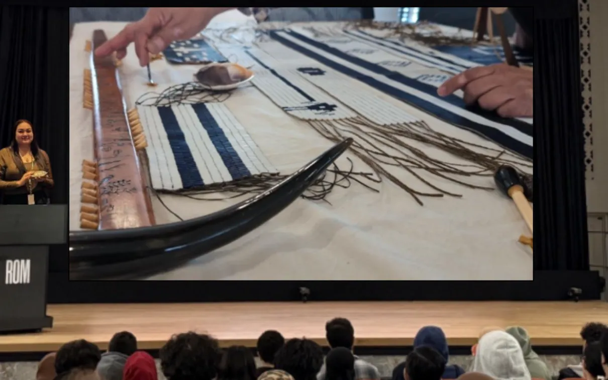 An Indigenous woman stands at a podium on a stage in front of a group of young people, gesturing to wampum belts and other belongings displayed on a screen.