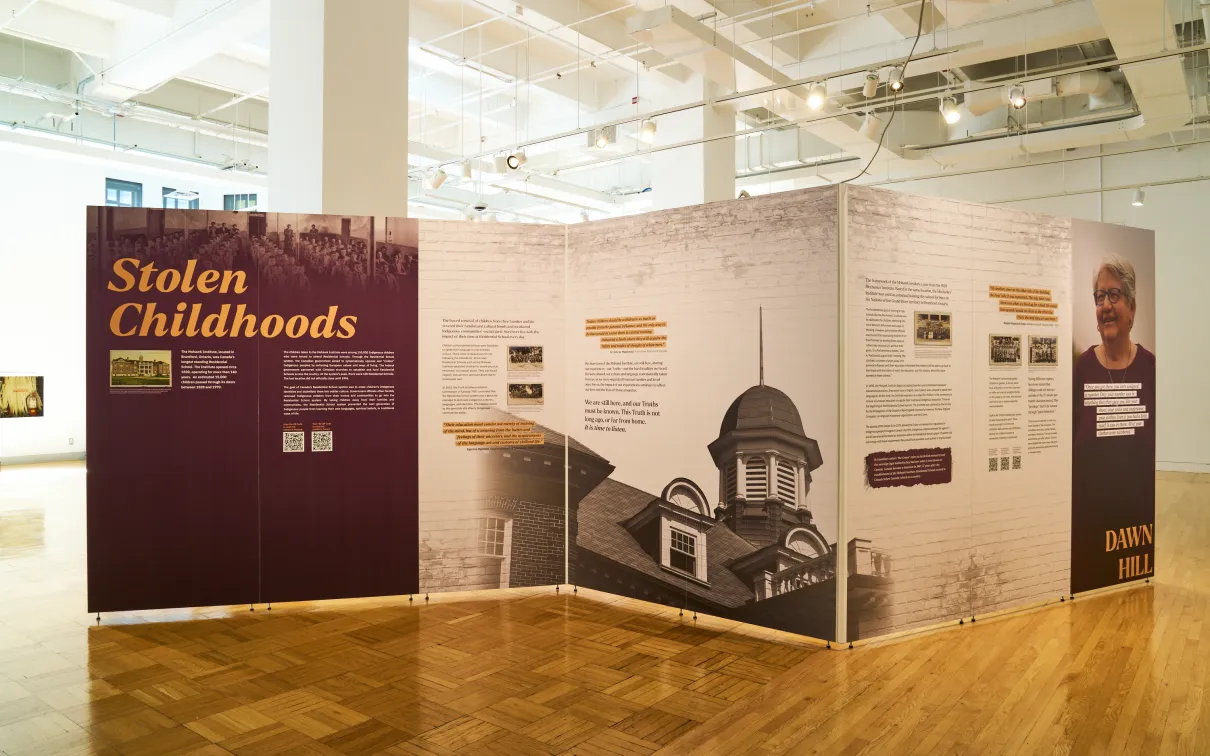 Large exhibition installation in a bright museum gallery featuring three angled display walls. The left panel, titled Stolen Childhoods, includes historic photographs and text about residential schools. The central panels display historical information, archival images, and a large black-and-white photograph of a school building. The right panel features a portrait and additional interpretive text. The exhibition stands on a polished parquet wood floor beneath high white ceilings with exposed lighting and d
