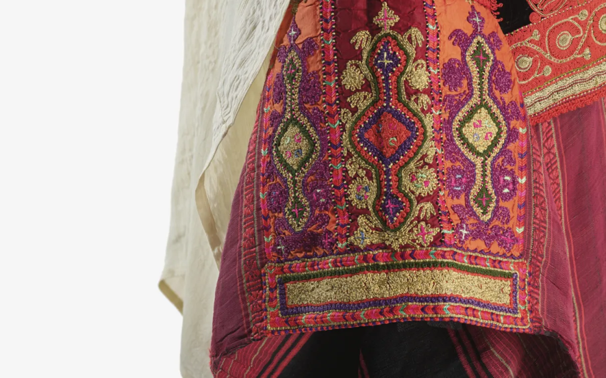 Close-up detail of a traditional garment featuring intricate red, orange, gold, and purple embroidery with geometric and floral patterns. A light-colored draped fabric falls alongside the richly textured textile, highlighting the craftsmanship and decorative stitching.