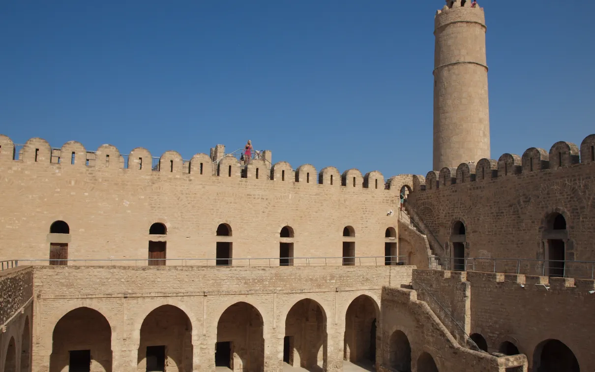 Alt text: An historic fortress with a tall, cylindrical tower under a clear blue sky.