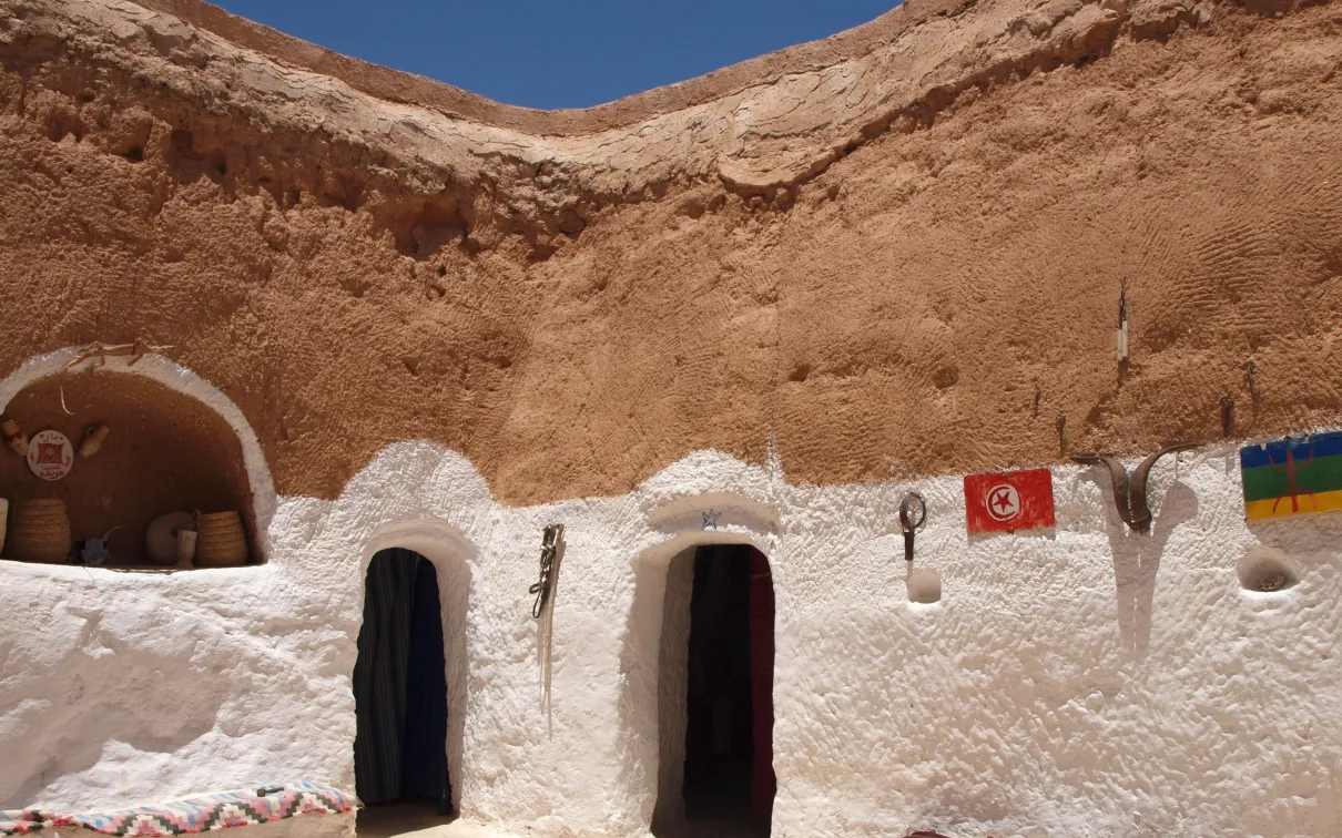 A picturesque, weathered white cave dwelling under a clear blue sky, with colourful flags and decorations on the wall.